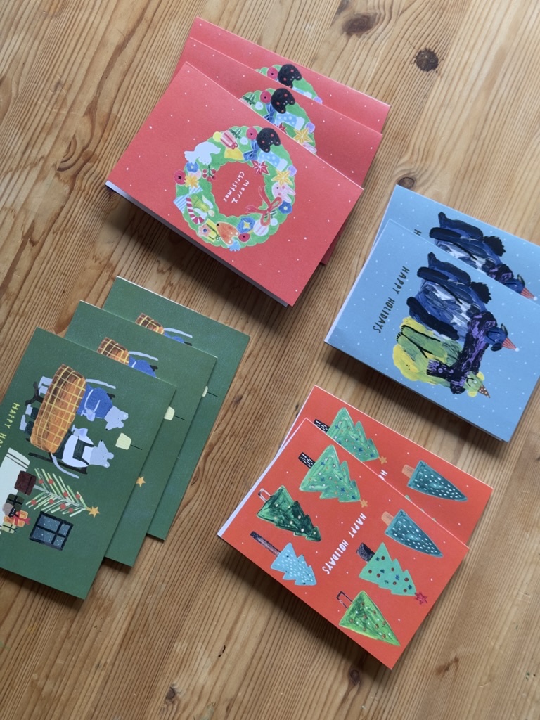 Ten illustrated Christmas and holiday greeting cards arranged on a wooden table.